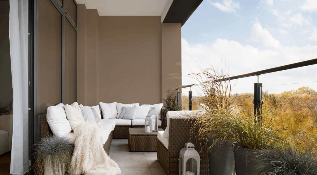 A contemporary balcony at the Kort View Condos | Luxury Living in Woodbridge is elegantly outfitted with wicker furniture and plush white cushions, complemented by a soft throw and lanterns. Potted plants adorn the glass railing, providing a picturesque view of trees under a partly cloudy sky.